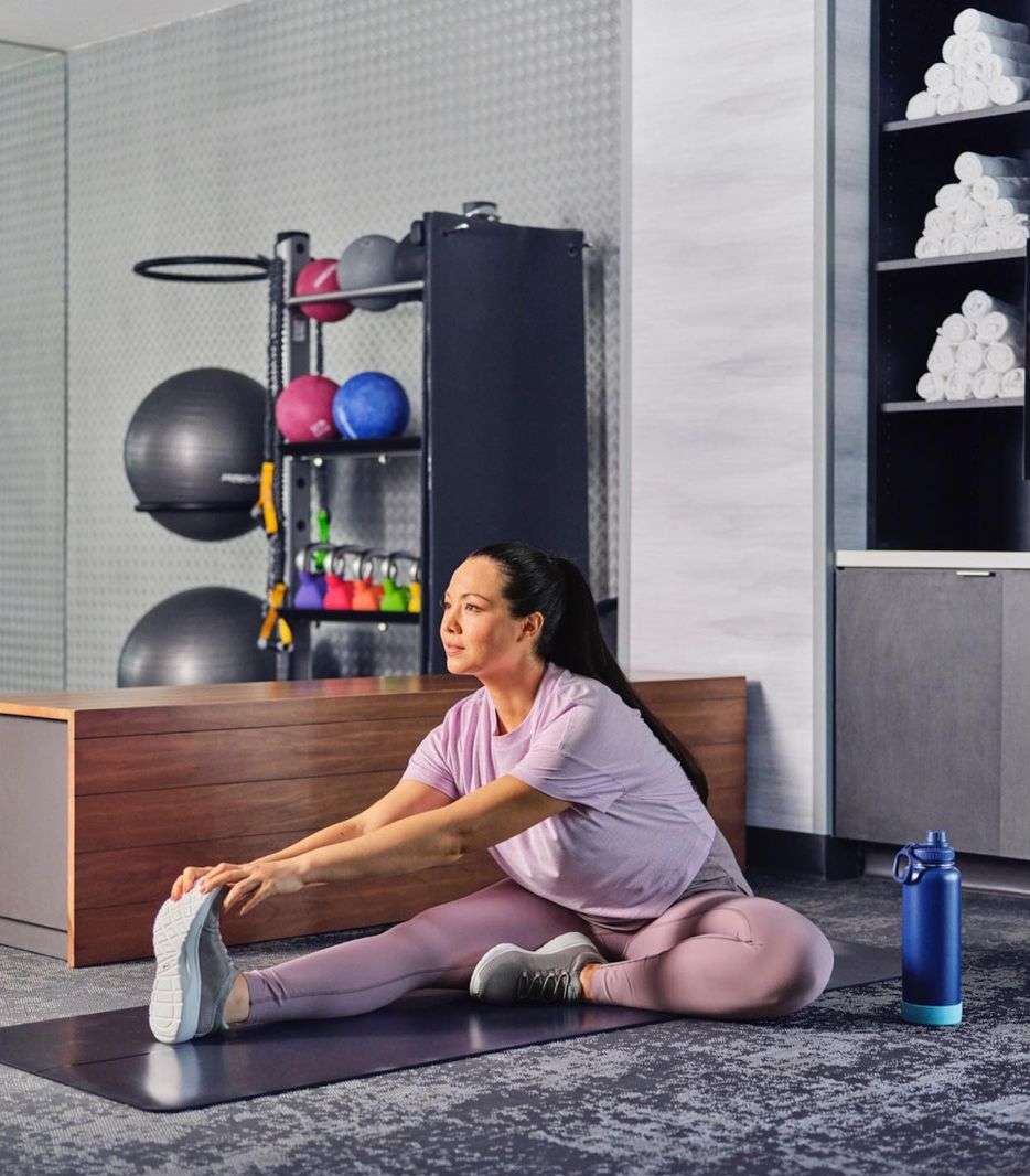 Athletic woman sitting on an exercise mat in a gym, stretching her leg in a full straddle split pose, wearing workout attire, indoors.
