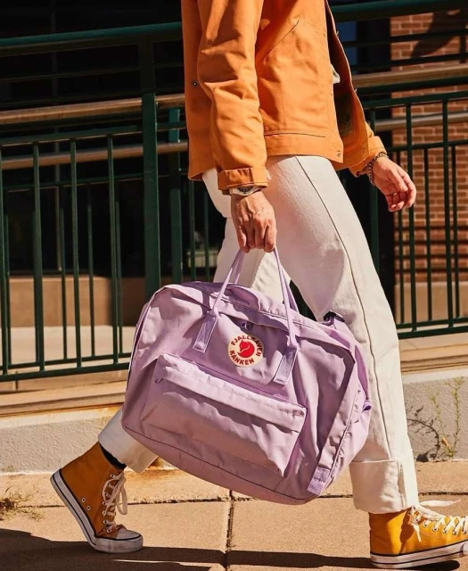 Person carrying a Fjallraven bag on a city sidewalk. Woman in orange jacket, white jeans, and Converse sneakers walks outside.