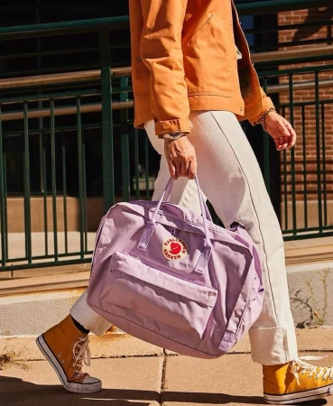Person carrying a Fjallraven bag on a city sidewalk. Woman in orange jacket, white jeans, and Converse sneakers walks outside.