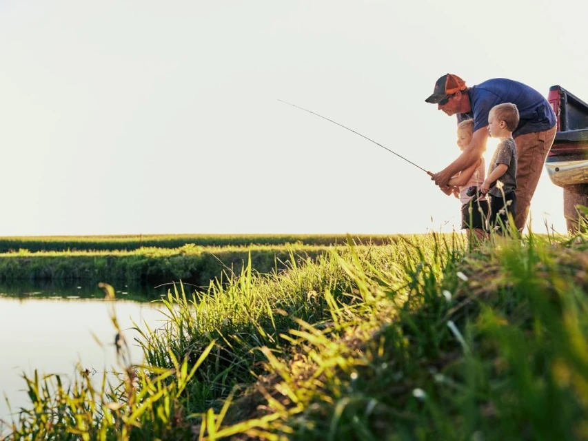 A father and two young children are fishing together near a pond surrounded by tall grass in a serene outdoor setting at sunset.