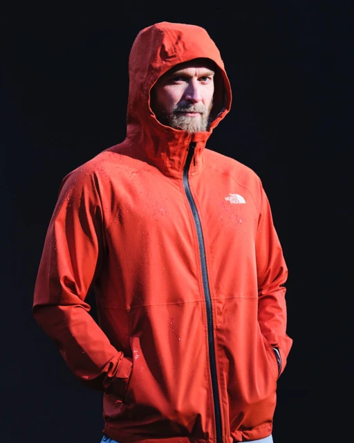 Man in orange hooded jacket stands against a dark background. Head and shoulders shot, facing slightly right, with water droplets on jacket.