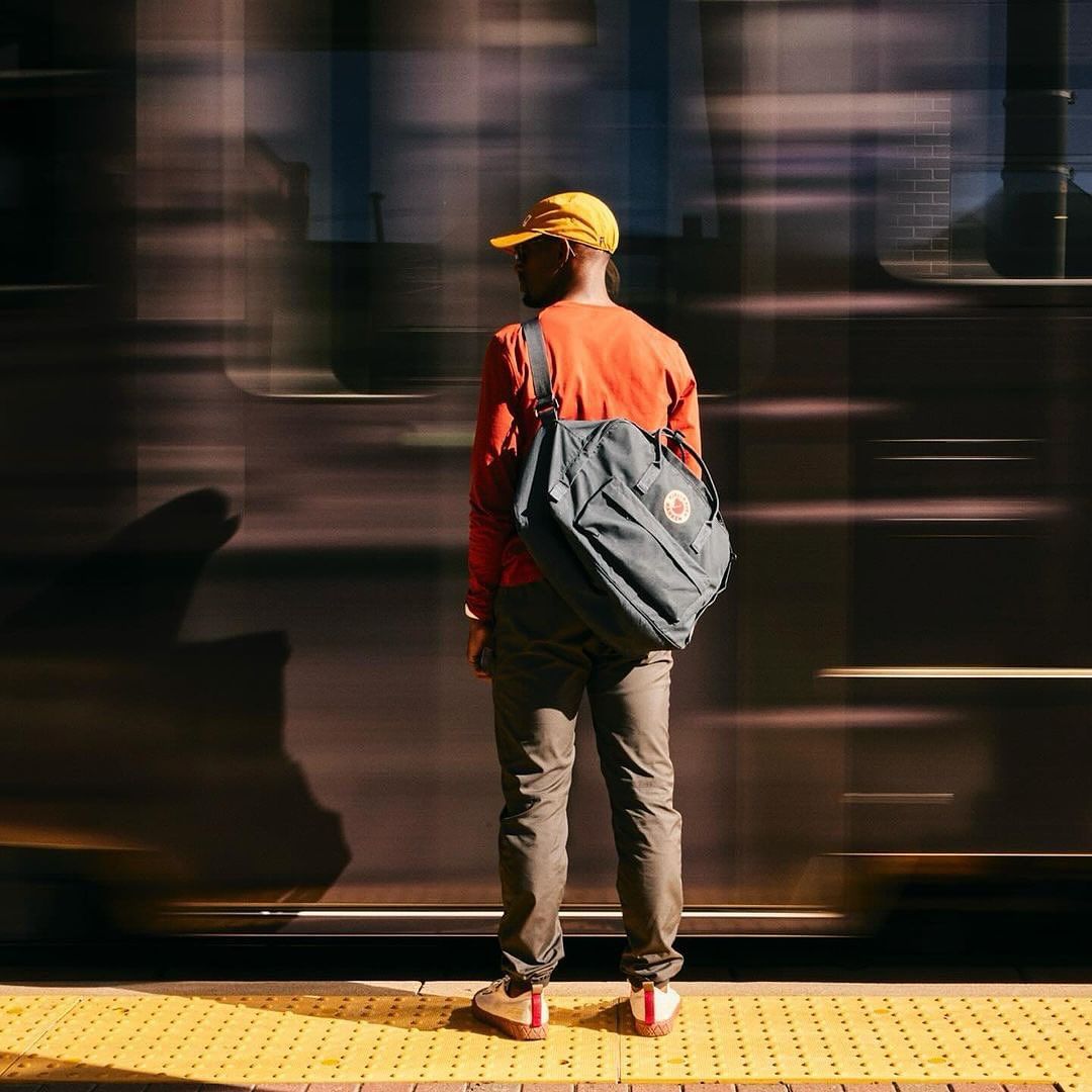 Back view of African-American man with backpack on train platform, wearing yellow cap, orange shirt, and standing on tactile paving.