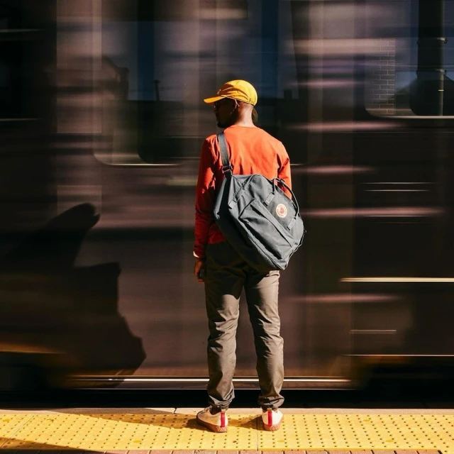 Back view of African-American man with backpack on train platform, wearing yellow cap, orange shirt, and standing on tactile paving.