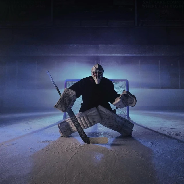 A hockey goalie in full gear positioned in front of a net on an ice rink, illuminated by bright, directional lighting with blue hues.