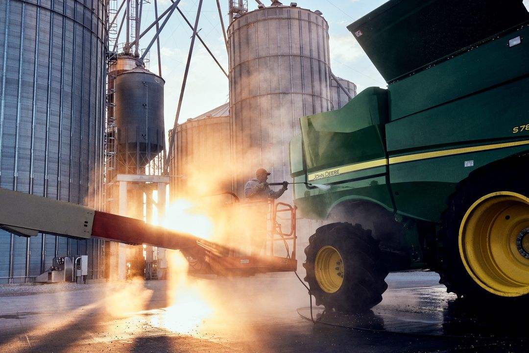 A person power washing a John Deere combine in front of grain silos, creating a sunlit haze in an outdoor agricultural setting.