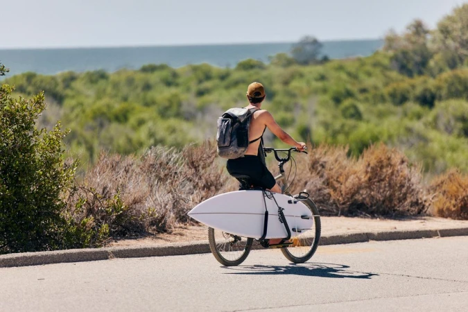 Back view of an adult on a bicycle transporting a surfboard on a paved road with a lush green and coastal background under a sunny sky.