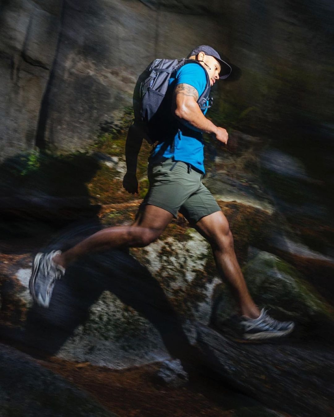 Athletic adult male trail runner wearing a backpack runs along a rocky hiking trail through a mountain landscape, illuminated by sunlight.