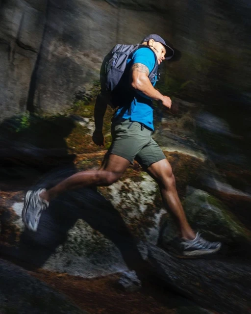 Athletic adult male trail runner wearing a backpack runs along a rocky hiking trail through a mountain landscape, illuminated by sunlight.