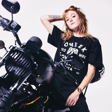 Red-haired woman wearing a black graphic t-shirt posing on a black motorcycle against a white backdrop.