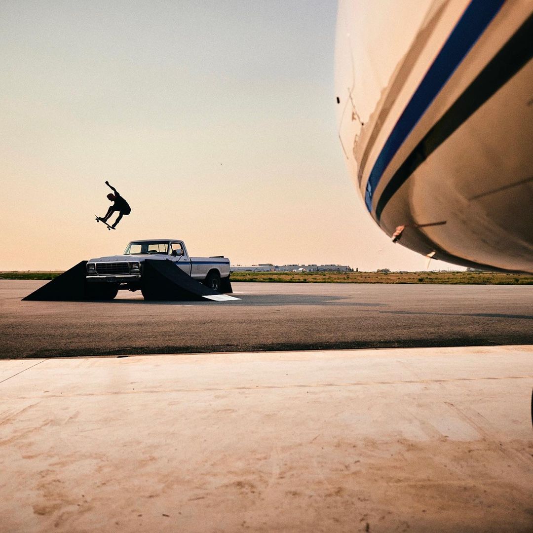 A skateboarder performs a trick over a ramp placed in front of a vintage pickup truck, with an airplane partially visible in the background.