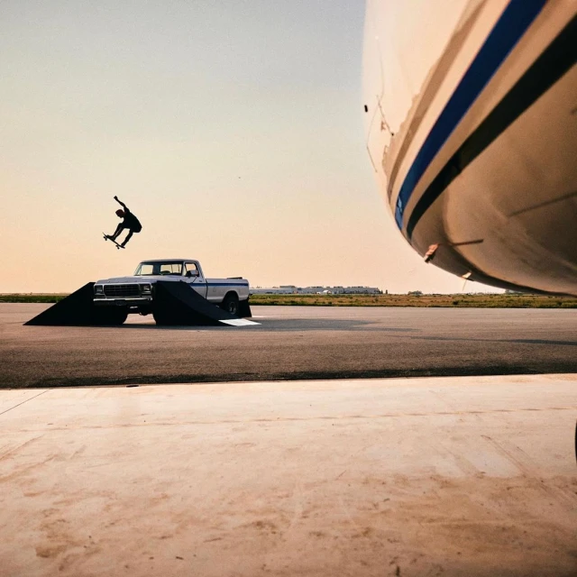 A skateboarder performs a trick over a ramp placed in front of a vintage pickup truck, with an airplane partially visible in the background.