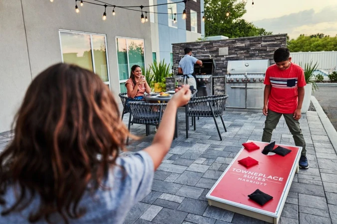 A group enjoys an outdoor game of cornhole on a patio with string lights above, while another person barbecues nearby at dusk.