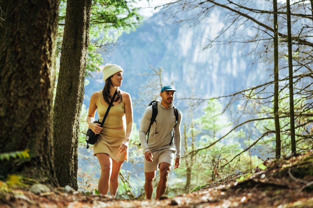 A couple hikes through a forest landscape with tall trees and a mountain backdrop, dressed in casual athletic wear in natural light.