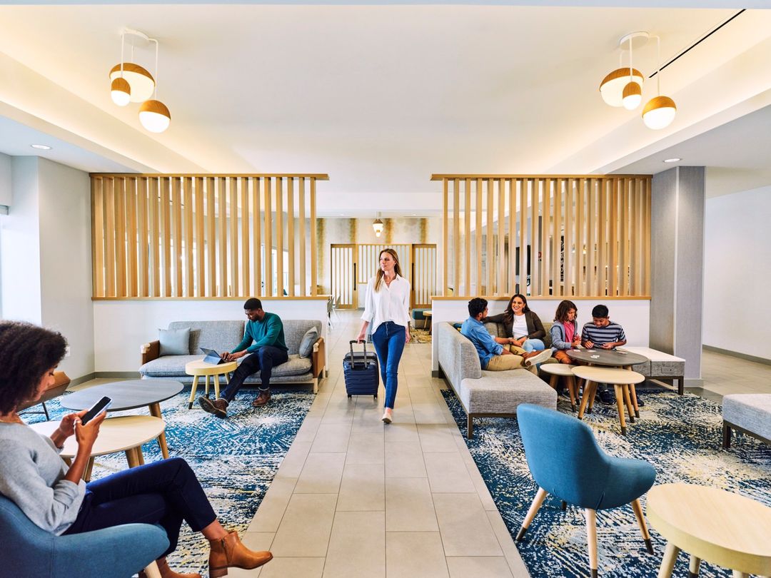 Modern hotel lobby with a diverse group of people relaxing on sofas and chairs; a young woman walks with a suitcase on wheels.