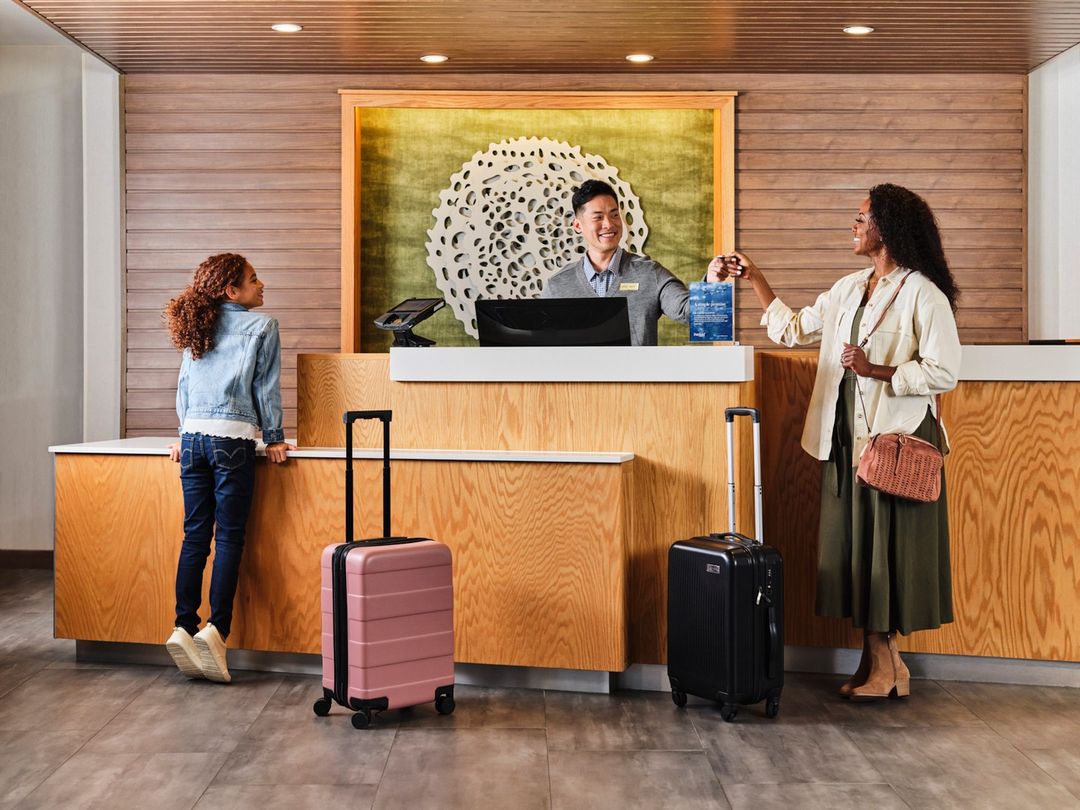 Woman checks in at hotel front desk with child; Asian concierge hands over the room key. Luggage sits on the floor.