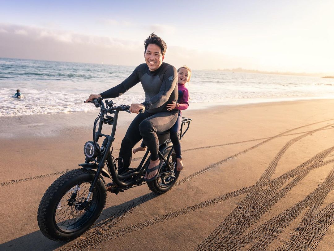 An adult male wearing a wetsuit smiles while riding an electric bicycle with a child on the beach in the late afternoon.