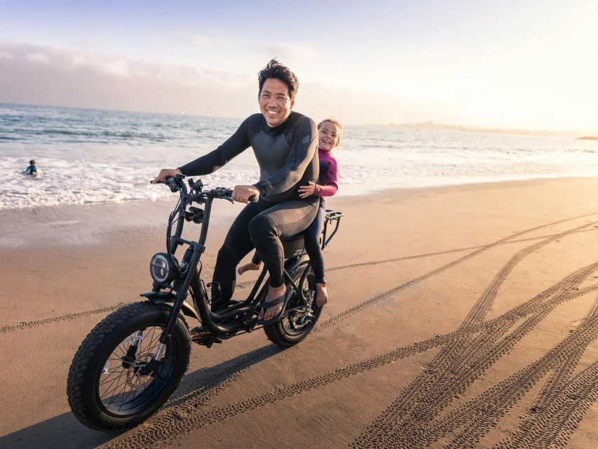 An adult male wearing a wetsuit smiles while riding an electric bicycle with a child on the beach in the late afternoon.