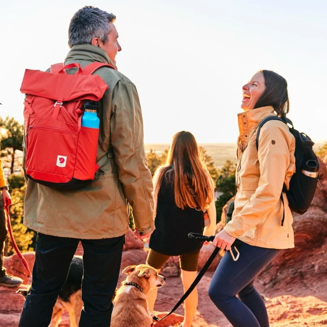 A group of four people hike on a red rock trail with two dogs. Two adults in the foreground, one with a red backpack, and one laughing.
