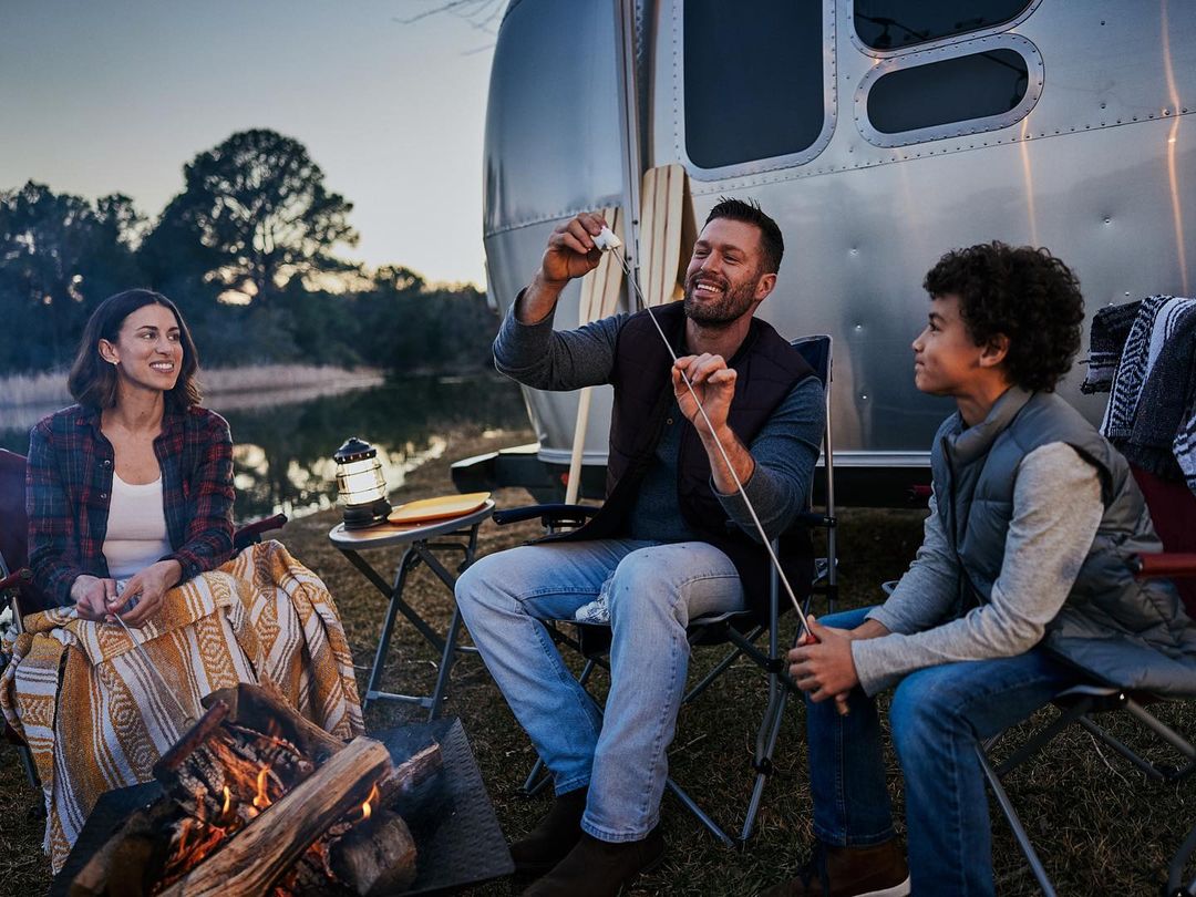 Family enjoys an outdoor campfire next to a silver Airstream trailer in the evening, roasting marshmallows on sticks near a lake.