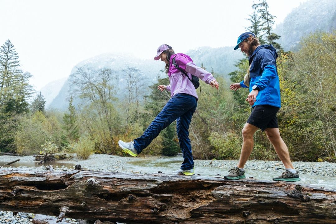Two hikers traverse a fallen tree over a stream in a misty forest. The woman is in purple, the man in blue hiking gear.