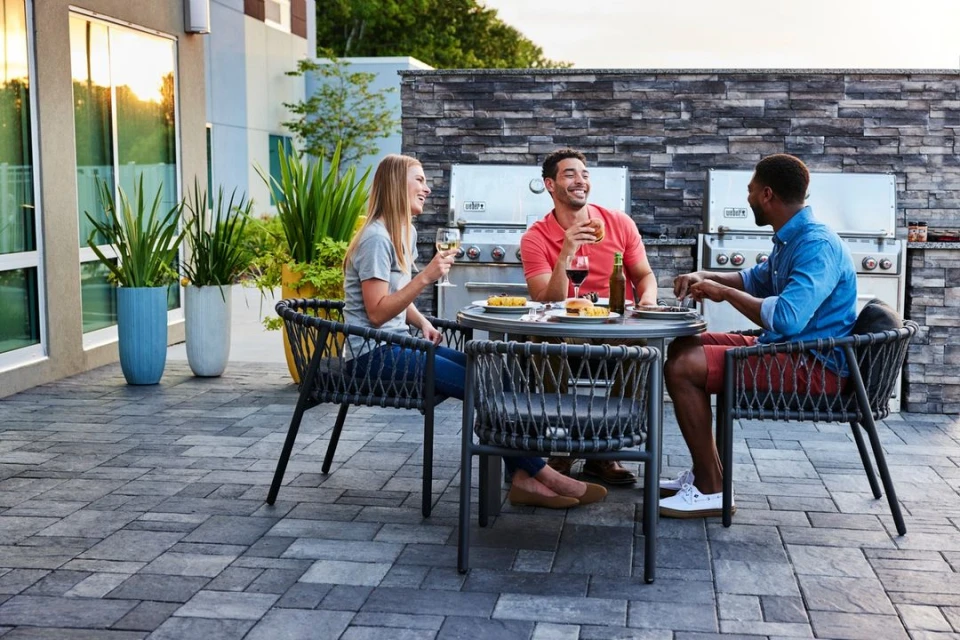 Three people sit at an outdoor table with food and drinks, surrounded by greenery and a stone wall with built-in grills at dusk.