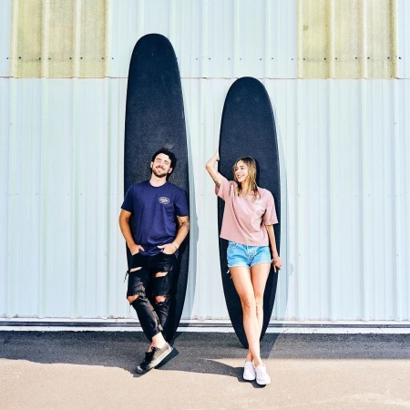 Man and woman standing outdoors against a corrugated metal wall, holding two black surfboards.