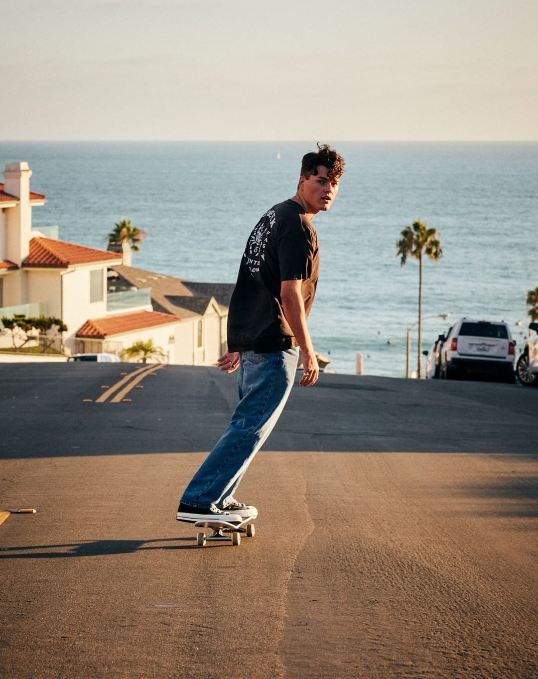 A young adult male is skateboarding downhill on an asphalt road near houses and palm trees, with the ocean in the distance.