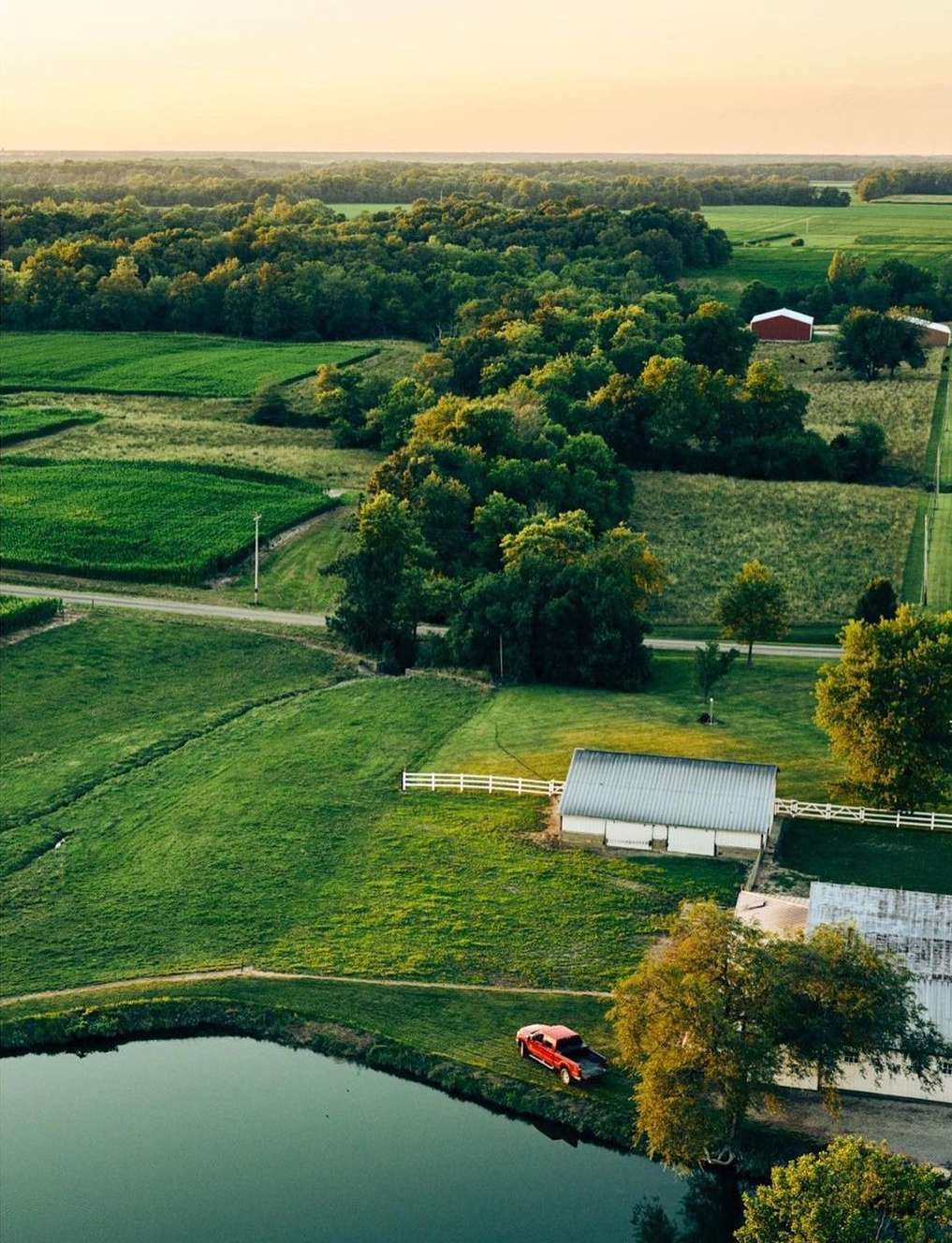 Aerial view of a rural landscape with farm buildings, green fields, a pond, trees, and a vintage red pickup truck.
