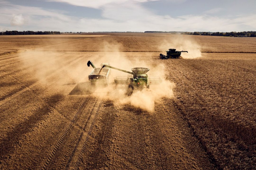 Aerial view of two combine harvesters working in a dusty agricultural field under a partly cloudy sky during the daytime harvest.