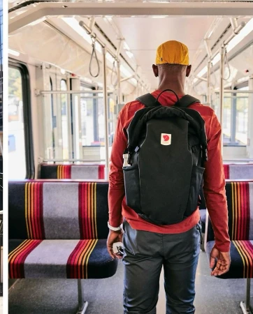 An individual with a backpack stands on a train with vibrant striped seats. The person wears a rust-colored shirt and a yellow hat.