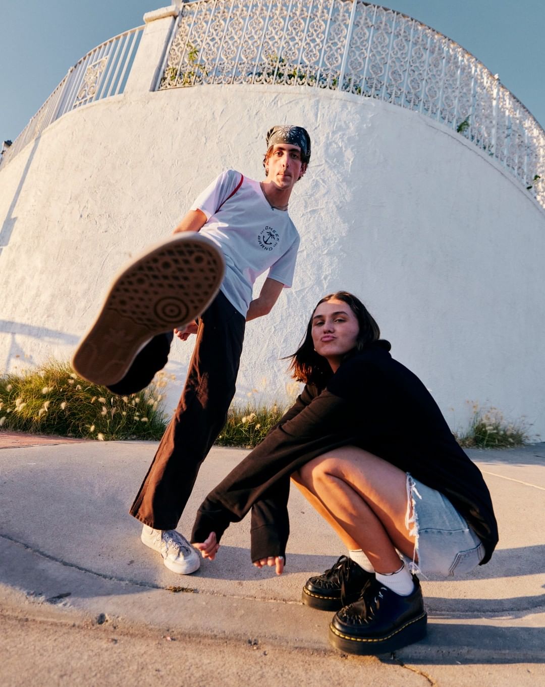 Young couple poses outdoors in front of white wall. Man in white shirt, brown pants with foot raised. Woman in denim shorts and black hoodie.