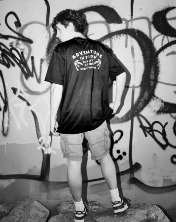 A young man seen from behind wearing a black graphic t-shirt and cargo shorts standing against a graffiti-covered wall.