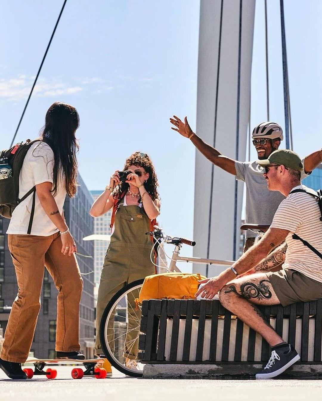 Group of four diverse friends enjoying an outdoor adventure on a bridge, featuring a skateboard, bicycle, and city views.