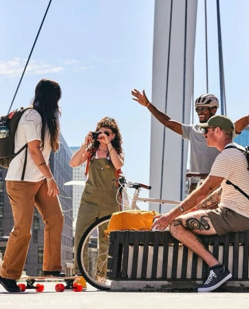 Group of four diverse friends enjoying an outdoor adventure on a bridge, featuring a skateboard, bicycle, and city views.