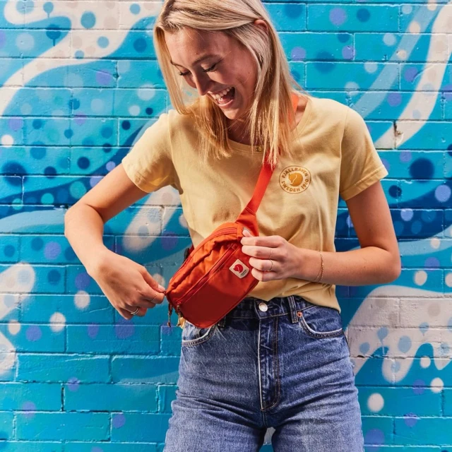 Blonde woman with a joyful expression zipping up an orange fanny pack worn across her body, with a patterned blue brick wall background.