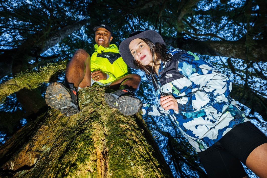 Two people, a man and a woman, sit on a tree branch wearing outdoor apparel in a forest with a low-angle view looking up at them.