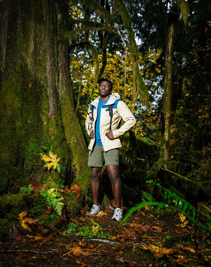 An adult man in outdoor attire stands next to a large mossy tree in a sunlit forest with green and yellow foliage.