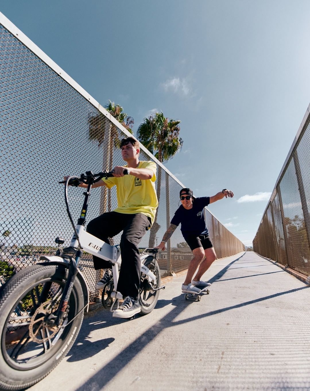 Two young men ride an electric bicycle and skateboard on an outdoor pedestrian bridge, surrounded by blue sky, metal fences, and palm trees.
