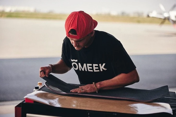 Man applies grip tape to a skateboard on a work bench outdoors near an airport runway. He wears a red cap and black T-shirt.