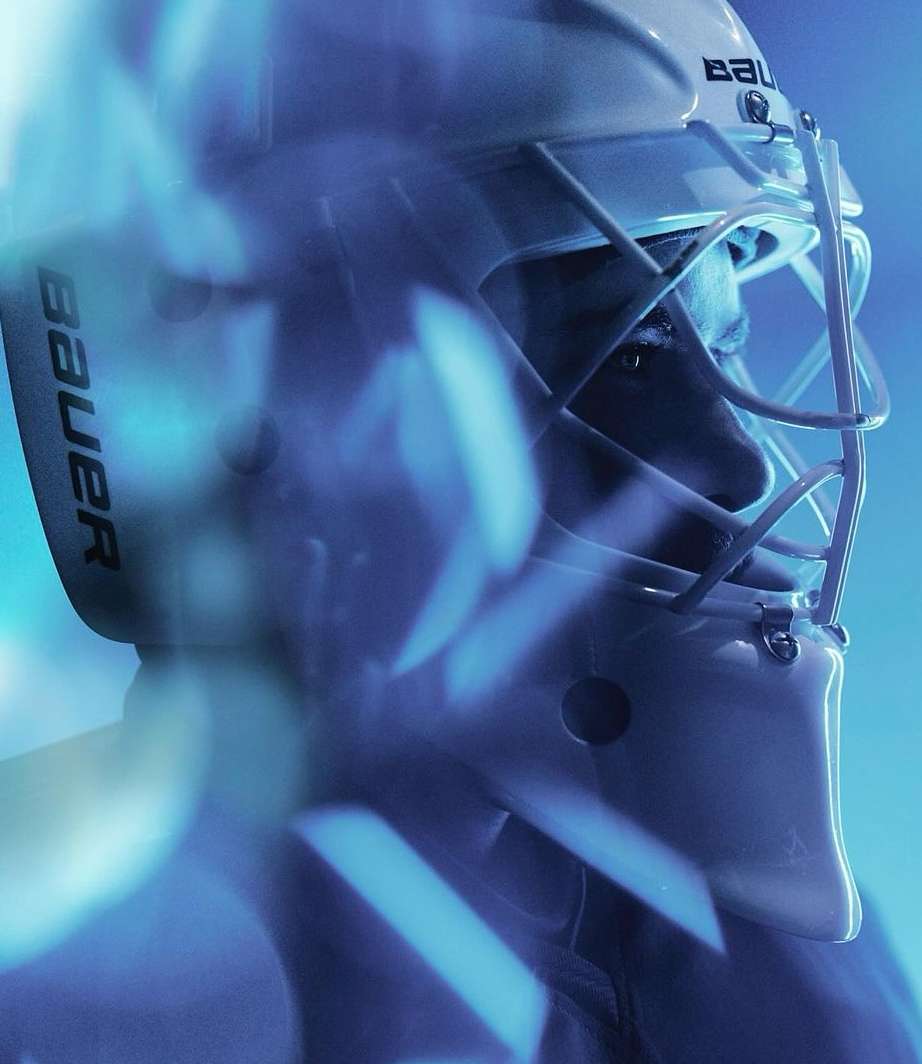 Close up of a hockey goaltender wearing protective Bauer mask and helmet, intense focus, blue lighting, sports photography.