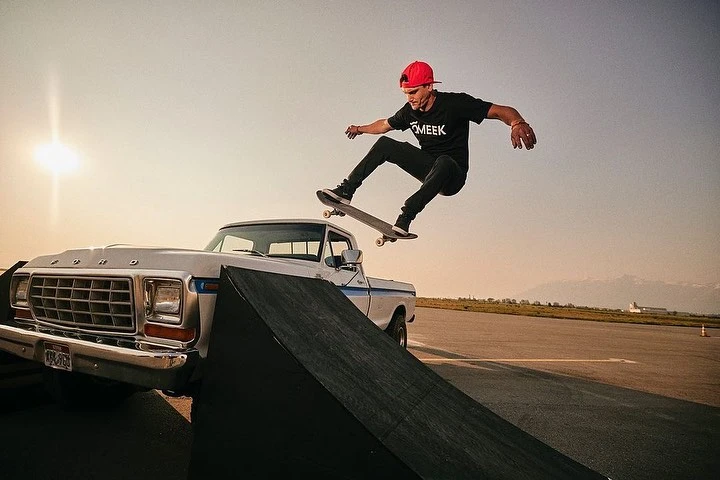 Man skateboarding off ramp in front of vintage truck at sunset. Red cap, black t-shirt, airborne on a skateboard, mountains in background.