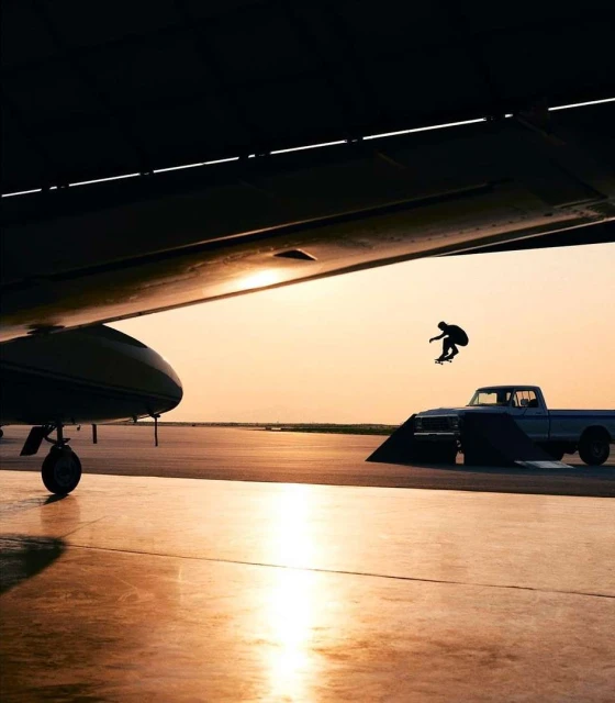 Skateboarder in mid-air performing a trick, silhouetted against a sunset, with an airplane wing and vintage truck in the background.
