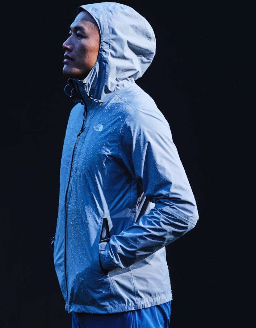 Profile view of a man in a grey hooded jacket standing against a dark background, featuring water droplets on the jacket's surface.
