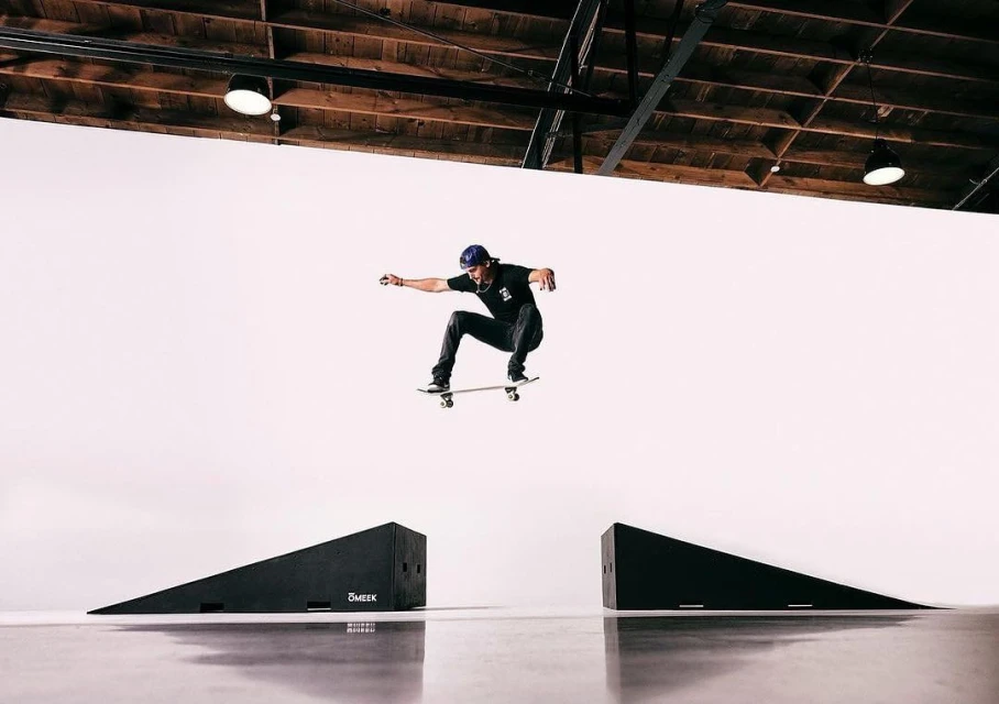 A male skateboarder in mid-air between two ramps inside a photo studio, performing a trick with arms outstretched and wearing a blue baseball cap.