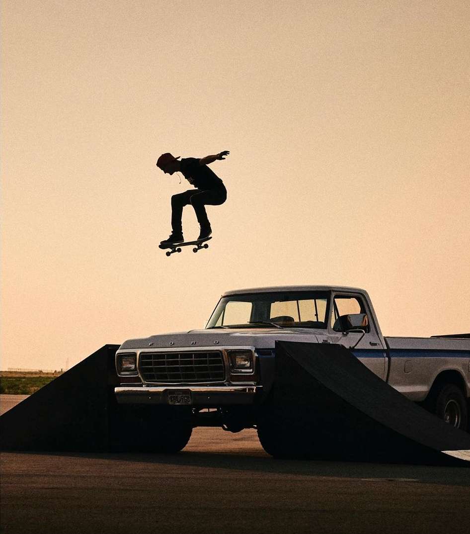 An adult in mid-air on a skateboard over a ramp and a vintage pickup truck against a soft sunset-colored sky, performing a stunt.