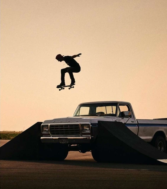 An adult in mid-air on a skateboard over a ramp and a vintage pickup truck against a soft sunset-colored sky, performing a stunt.