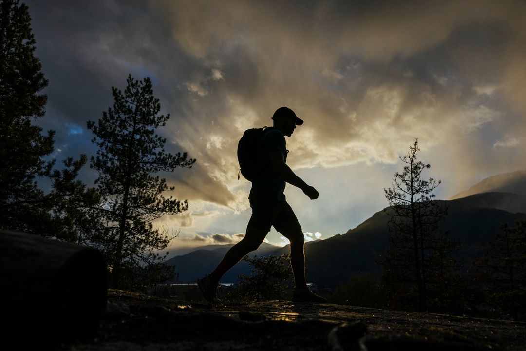 Silhouette of a man with a backpack walking through a forest landscape at dusk with a dramatic cloudy sky and sun rays.