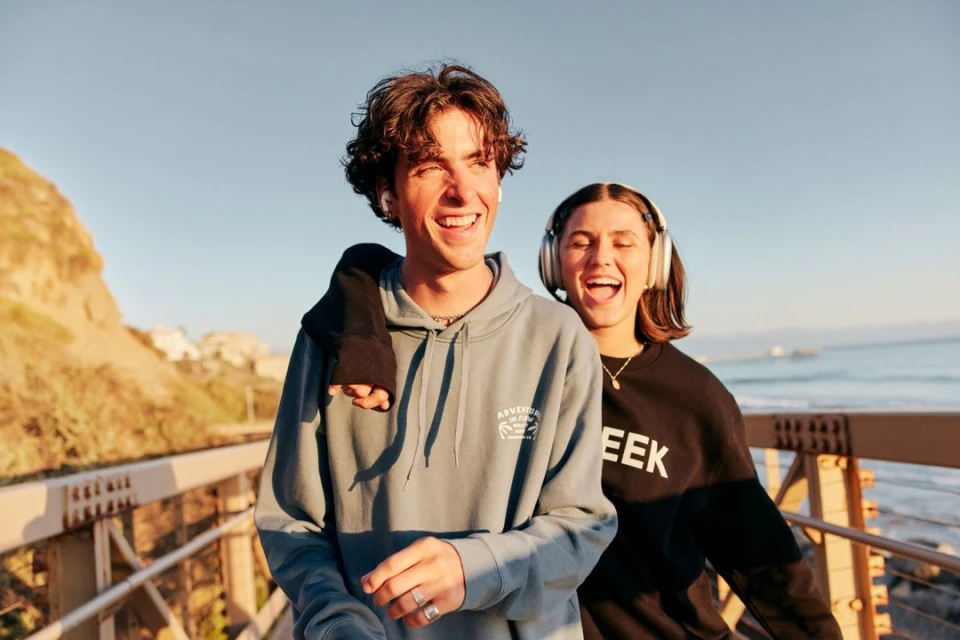 A young couple, smiling and embracing on a pathway with a blue sky and ocean view. They are wearing casual clothing and headphones.