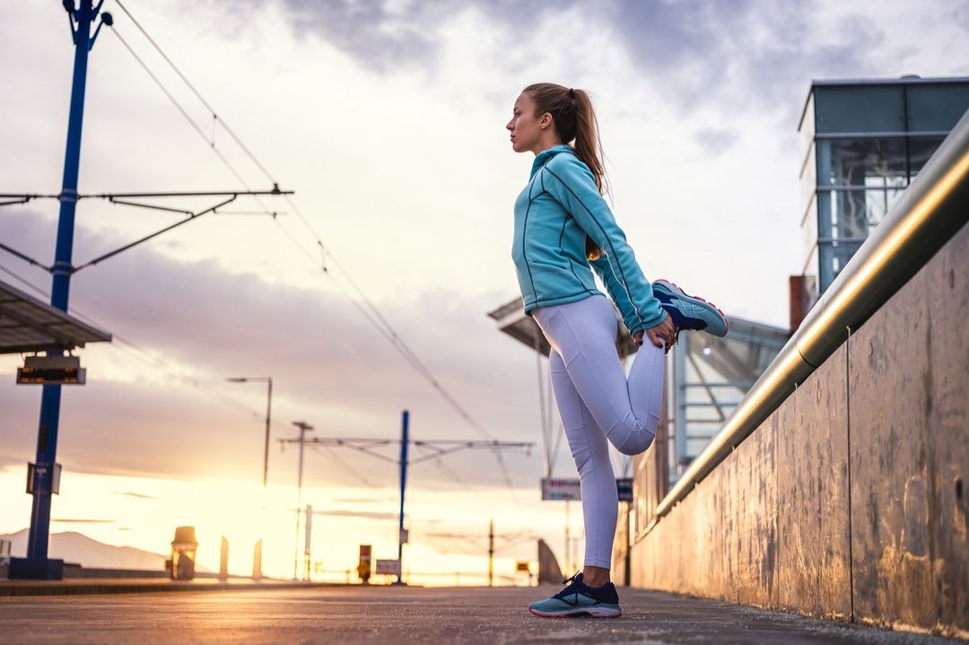 Athletic woman in sportswear stretches her leg outdoors during sunrise, with urban infrastructure and cloudy sky in the background.