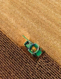 Aerial view of a green combine harvester operating in a vast golden agricultural field, capturing rows of harvested crops.